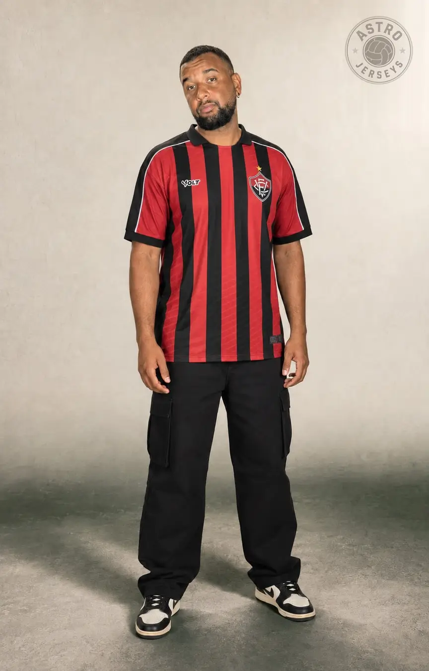 Pessoa de corpo inteiro vestindo a camisa Vitória 26/27, rubro-negra, em estúdio de fundo neutro